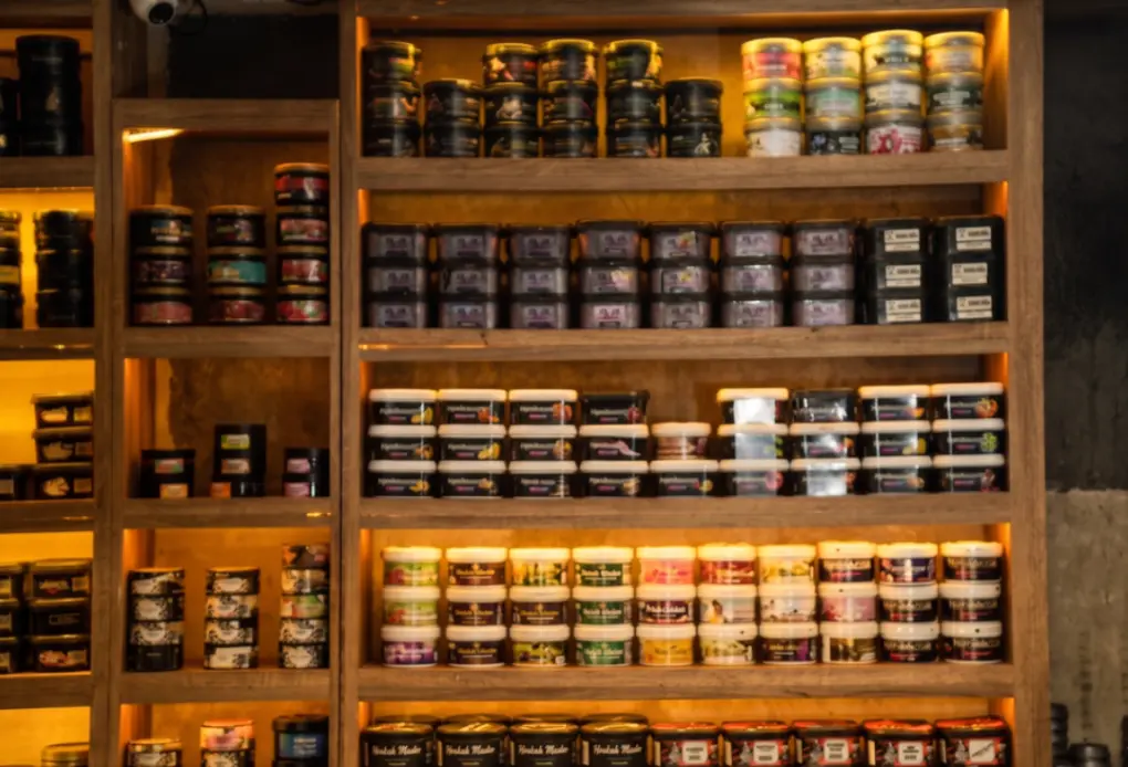 A warmly lit, extensive display of stacked premium shisha tobacco containers from brands like Tombacco, organized neatly on retail shelves, inviting customers to buy shisha in Bali.