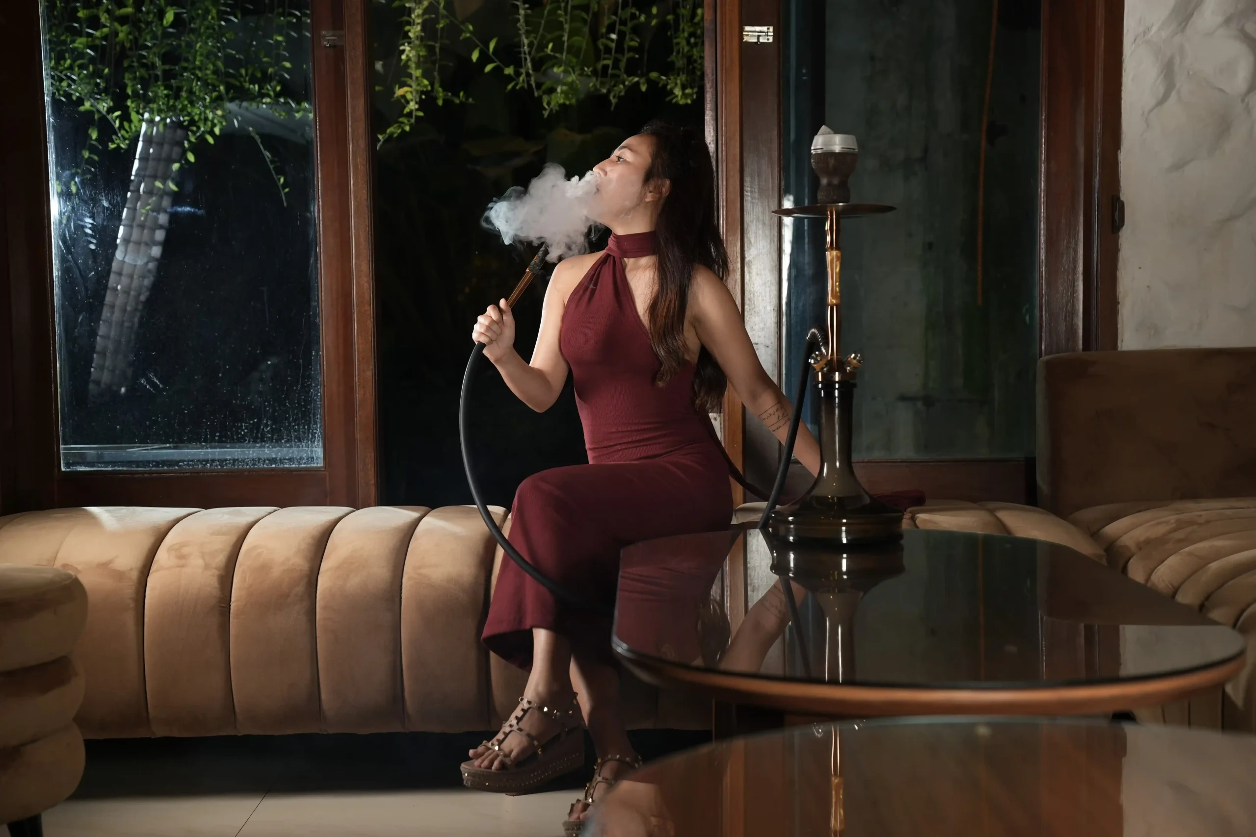 A woman in an elegant red dress relaxing on a cozy sofa, blowing a thick cloud of smoke while enjoying her hookah bali at home.