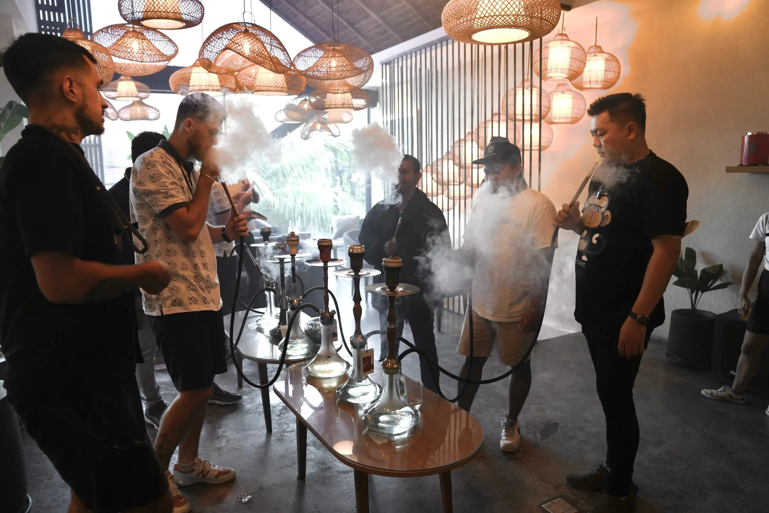 A group of friends having a great time hanging out indoors, standing around a table with several devices during a fun social hookah bali gathering.