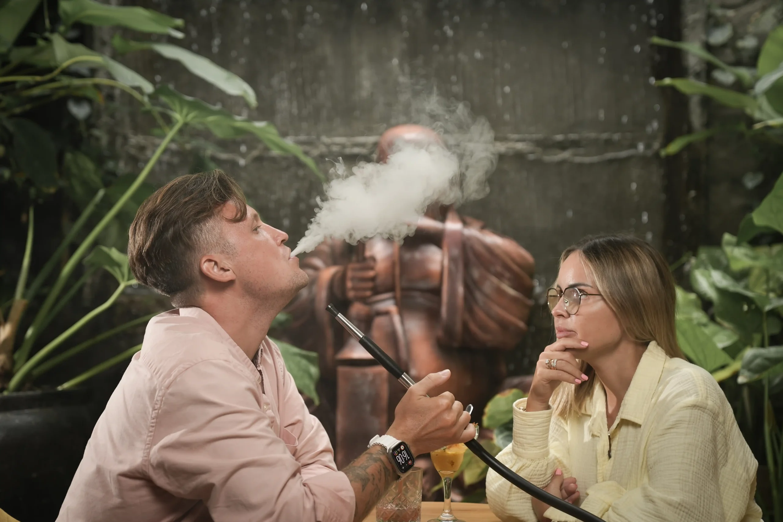 A man gracefully blowing a thick cloud of shisha smoke while relaxing with a woman in a lush, green, outdoor lounge setting. A perfect representation of enjoying premium Adalya flavor alternatives with a quality home or cafe setup.